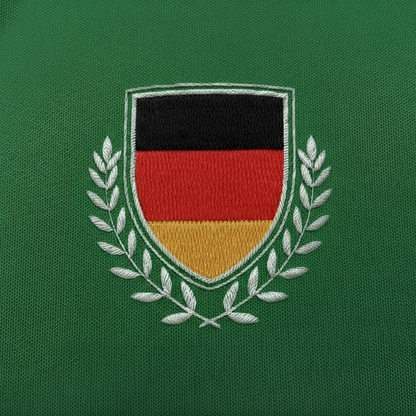 Detailaufnahme der Vorderseite des grünen Fan-Polo Deutschland 2026 mit gesticktem Deutschland-Wappen in Schwarz-Rot-Gold und weißen Ölzweigen